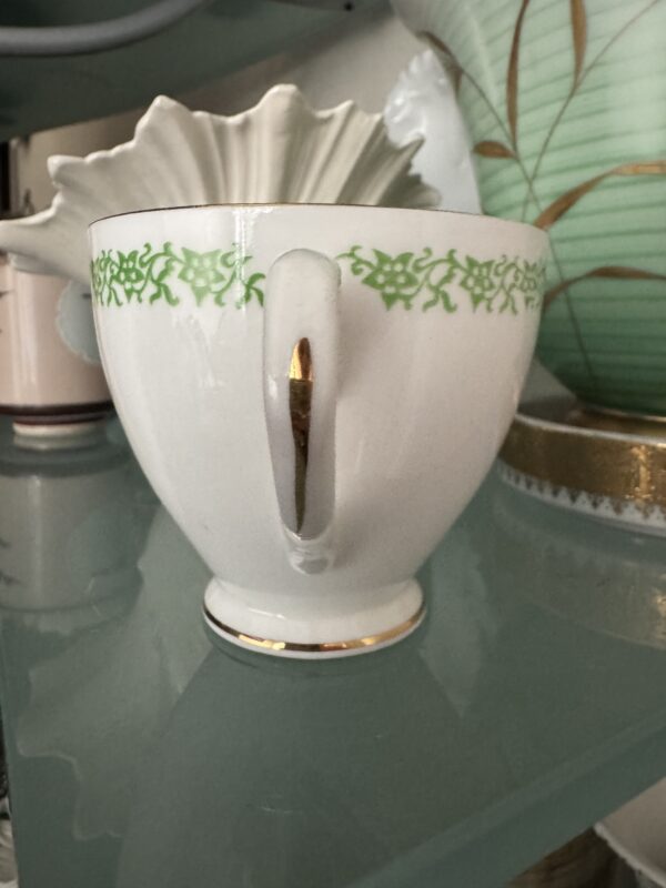 The English Ivy Teacup, a white porcelain cup with a gold rim and handle, features a band of green English Ivy near the rim. It sits on a glass shelf with decorative pottery and dishes in the background.