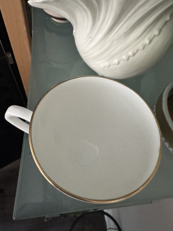 The English Ivy Teacup, featuring a white body and gold rim, rests on a glass shelf. Soft lighting highlights its elegant design, complemented by a decorative ceramic piece and the subtle charm of English Ivy in the background.