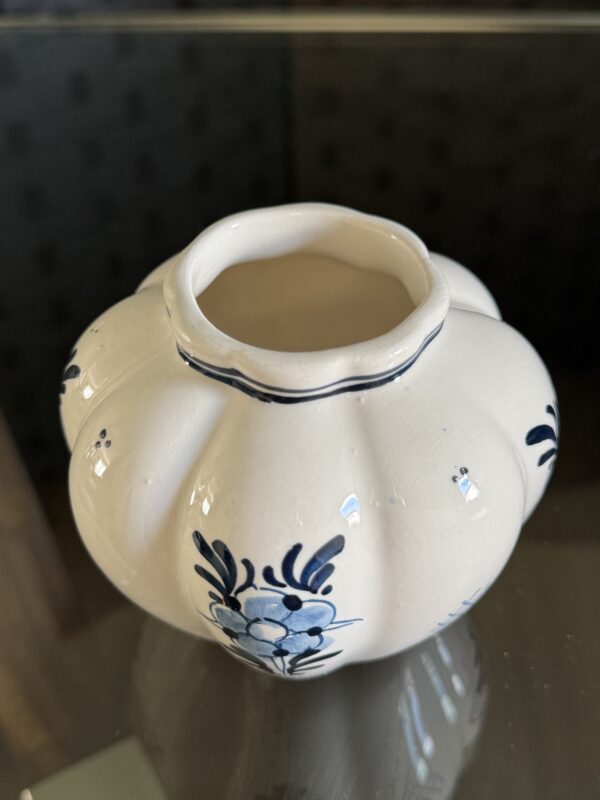 The English Ivy Teacup is a small, white ceramic cup with a scalloped rim and ribbed body. Blue, hand-painted floral designs wrap around it, evoking English ivy. It rests on a reflective glass surface against a dark background.