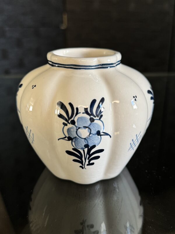 The English Ivy Teacup is a small, round, cream ceramic vase with a scalloped teacup shape, featuring blue floral designs and thin blue lines. It is displayed on a reflective surface with a dark, blurred background.