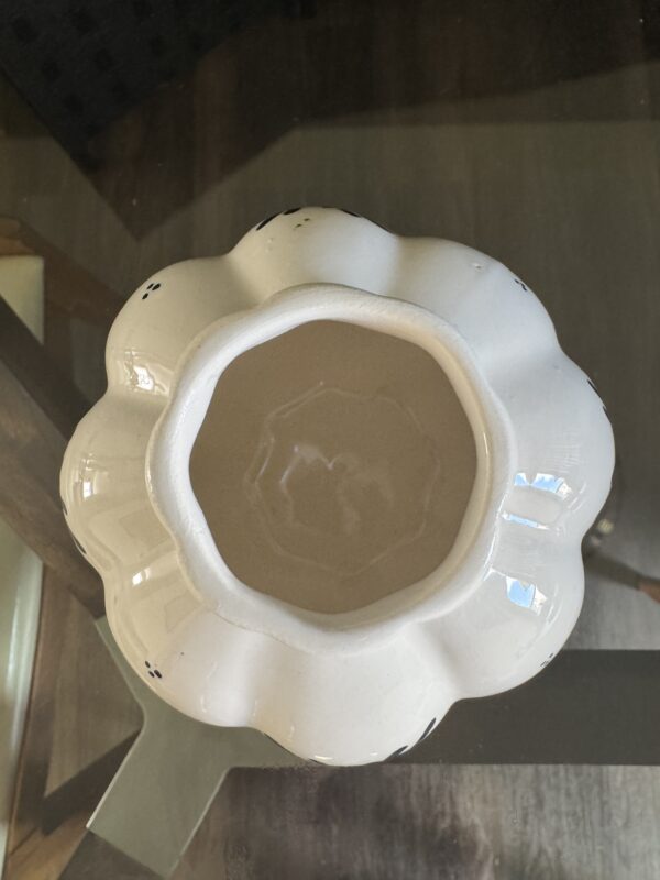 The English Ivy Teacup, crafted from white ceramic with a wavy, scalloped rim and beige interior, sits atop a glass tabletop. Glossy reflections highlight natural light, with nearby wooden furniture and an ivy leaf adding charm.