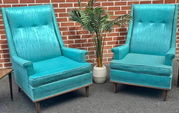 The Kroehler Wingback Chair Set, upholstered in retro turquoise with wooden legs, is arranged on grey pavement beside a leafy potted plant, set against a red brick wall for a classic mid-century modern look.