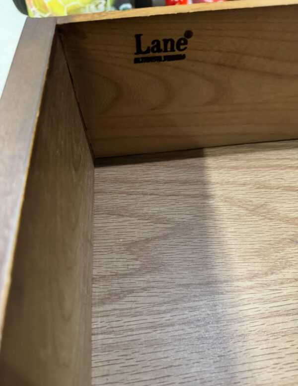Close-up of the inside of a Lane Side Tables, Pair drawer. The wood features a light natural finish and a black-stamped Lane® Altavista, Virginia logo on the back panel. Colorful objects rest on the countertop visible at the top edge.