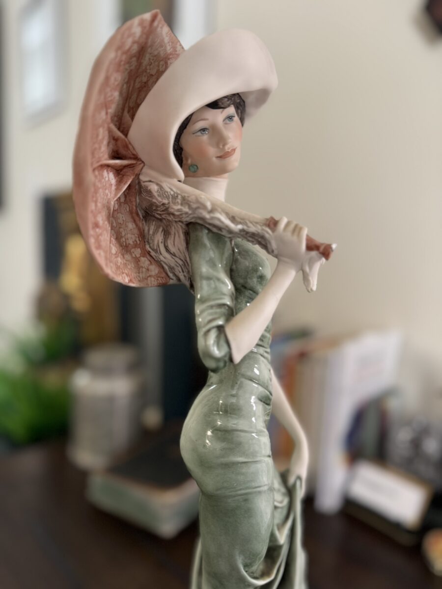 The Armani "After Noon" Figurine depicts a woman in a green dress and pink hat, holding a matching parasol over her shoulder. She stands on a table with books and blurred decor in the background.