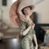 The Armani "After Noon" Figurine depicts a woman in a vintage green dress and wide-brimmed hat, holding a pink parasol over her shoulder, with an elegant background suggesting a staircase with books and furniture.