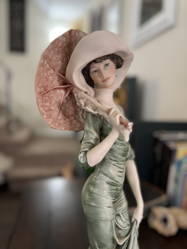 The Armani "After Noon" Figurine depicts a woman in a vintage green dress and wide-brimmed hat, holding a pink parasol over her shoulder, with an elegant background suggesting a staircase with books and furniture.