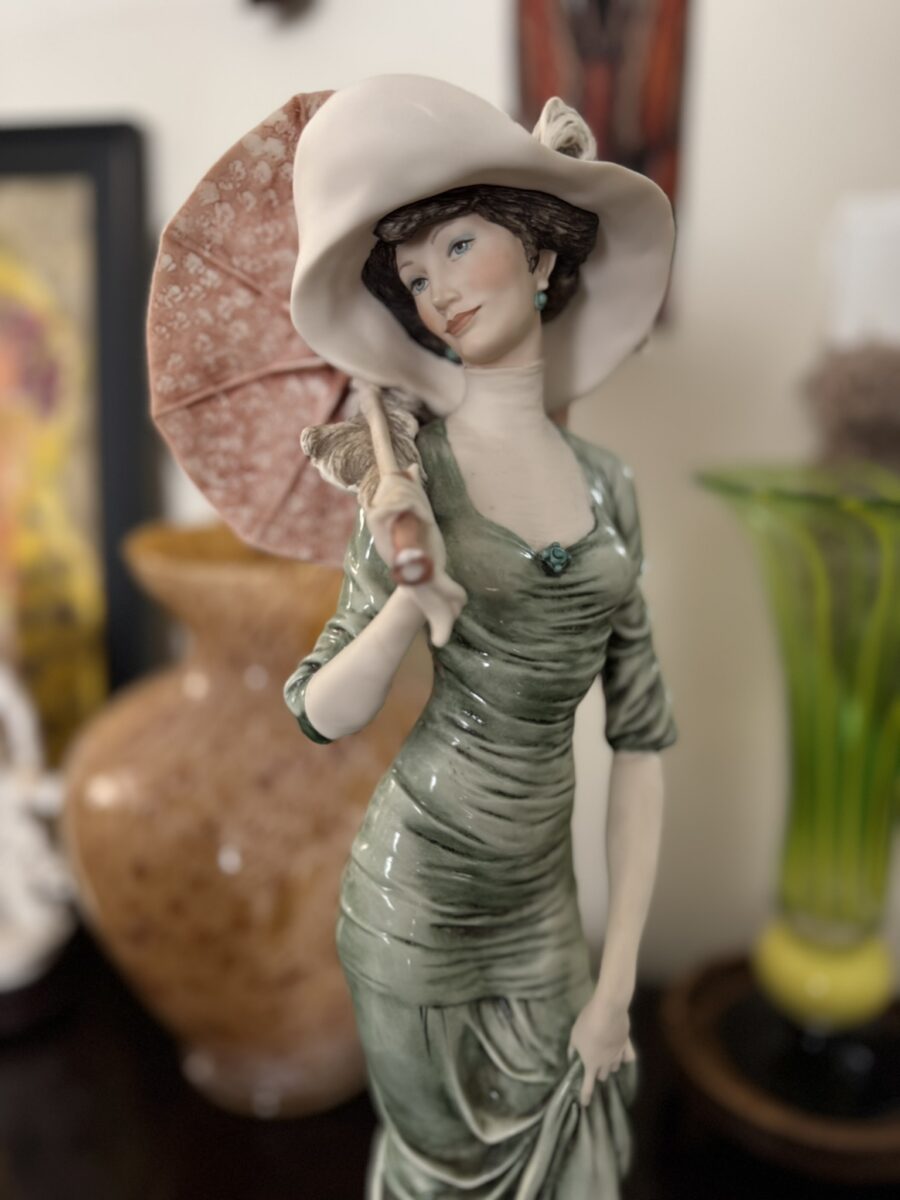 The Armani "After Noon" Figurine depicts a woman in a green dress and wide hat holding a pink parasol, with short brown hair and a calm look—an elegant addition to any vintage-inspired decor.