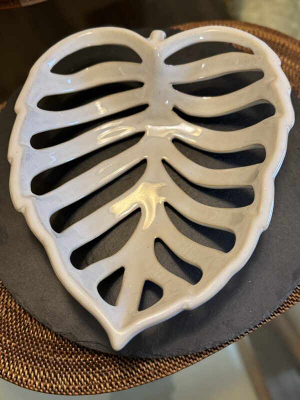 A glossy white ceramic monstera leaf dish sits on a dark slate plate and brown placemat, its abstract cut-outs echoing the artistic flair of the Eastern Art Glass Abstract Vase.