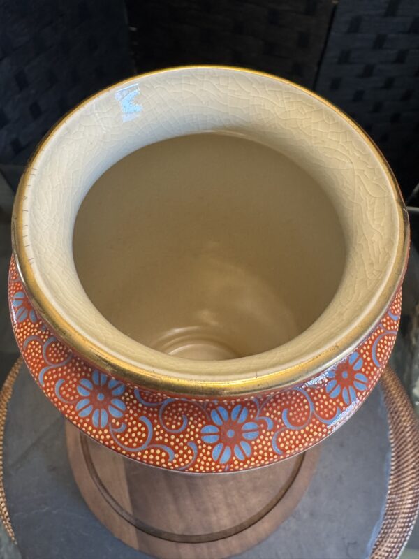 The Eastern Art Glass Abstract Vase features a wide, round opening, cream interior, and rim adorned with red and orange dots alongside blue flowers. Displayed on wood against a dark textured background.