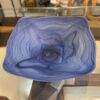 A Blue Swirl Art Glass Bowl, square-shaped with gently undulating edges and swirling deep blue patterns, rests on a glass table. Reflections and shelves with decorative objects appear in the softly blurred background.