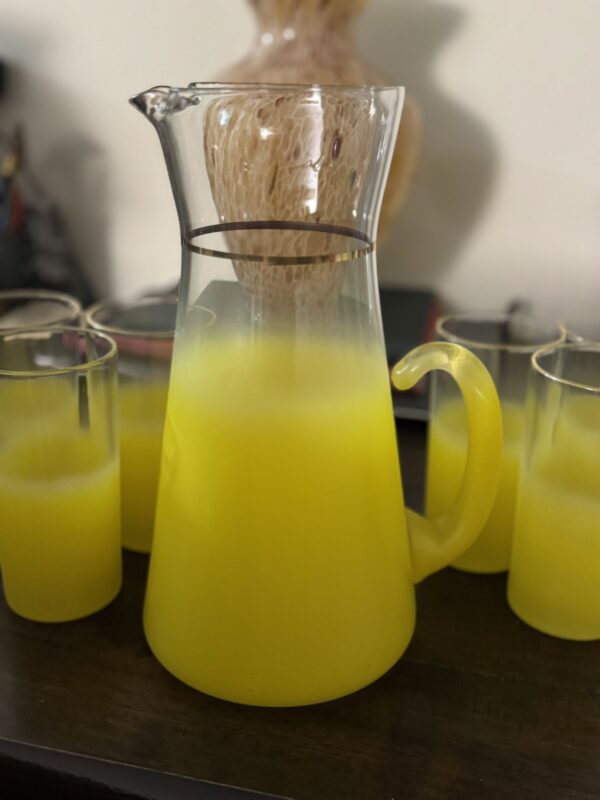 A large glass pitcher with a yellow handle sits on a dark wooden surface, filled with bright juice and surrounded by matching glasses. In the background, the Eastern Art Glass Abstract Vase adds artistic flair to the scene.