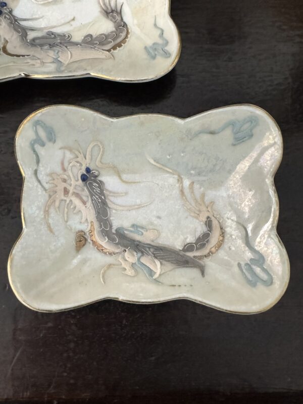 The Chinese Lidded Tea Cup features a painted dragon in shades of gray, blue, and tan on a pale, glossy surface with stylized blue clouds and gold-trimmed rim, embodying elegant Chinese design.