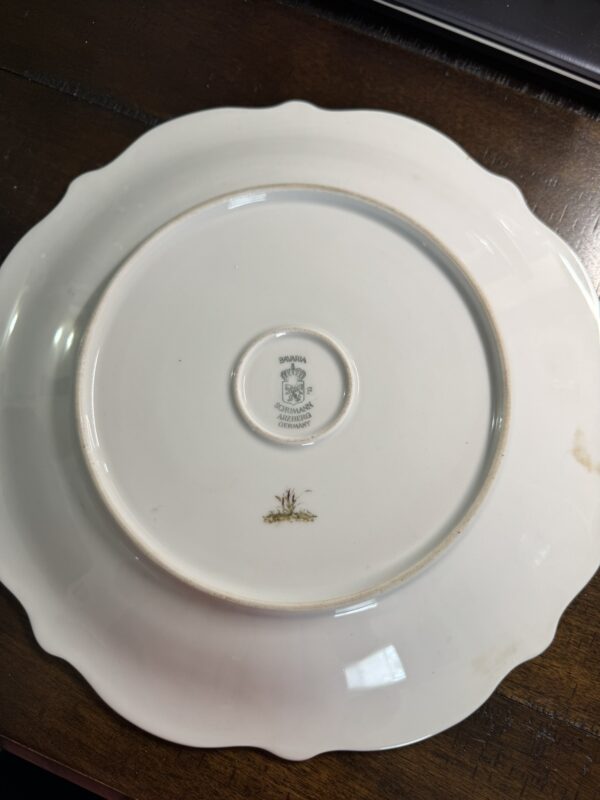 A white ceramic plate with scalloped edges is upside down on a dark wooden surface next to a Blue Crackle Glass Vase. The plate’s center shows two markings: a greenish emblem with text and a smaller gold design.