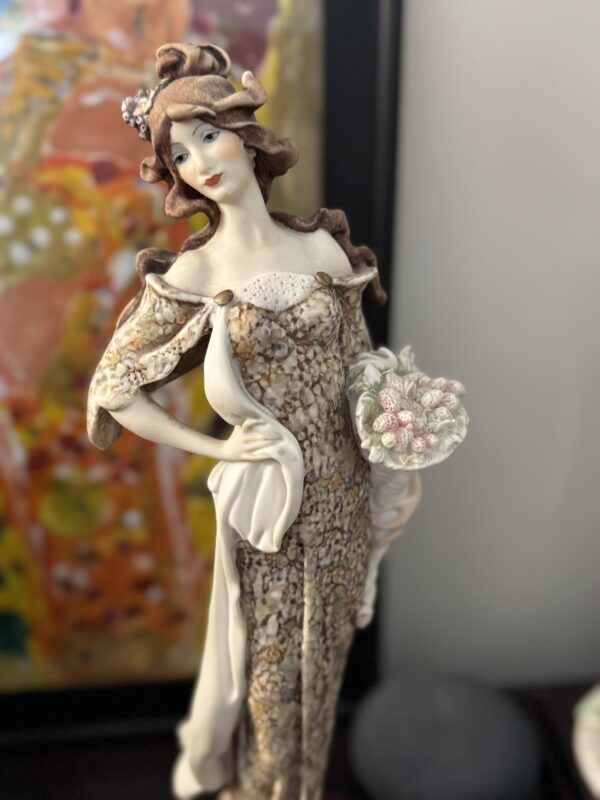 The Armani Lady with Strawberries Figurine features a porcelain lady in a vintage-style dress, holding strawberries. She has curled brown hair adorned with small flowers and poses with one hand on her hip, set against a softly blurred abstract painting backdrop.