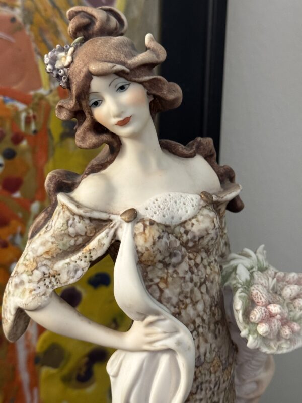 A close-up of the Armani Lady with Strawberries Figurine, featuring wavy brown hair and an ornate marbled dress, holding a basket of strawberries and set before a blurred, colorful abstract background.
