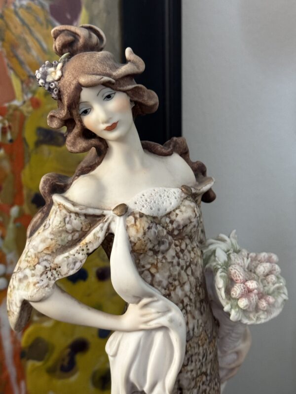 The Armani Lady with Strawberries Figurine features a delicately painted woman with wavy brown hair, holding strawberries and wearing an elegant flowing off-the-shoulder dress, her gentle smile set against a colorful backdrop.