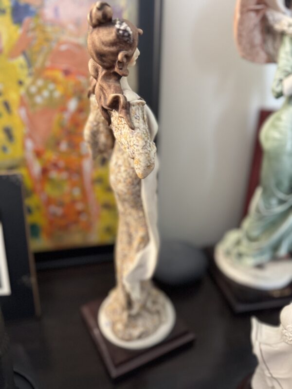 The Armani Lady with Strawberries Figurine features a ceramic lady with an ornate updo atop a round base, dressed in a textured, flowing robe with long sleeves. A colorful abstract painting and other sculptures appear in the background.