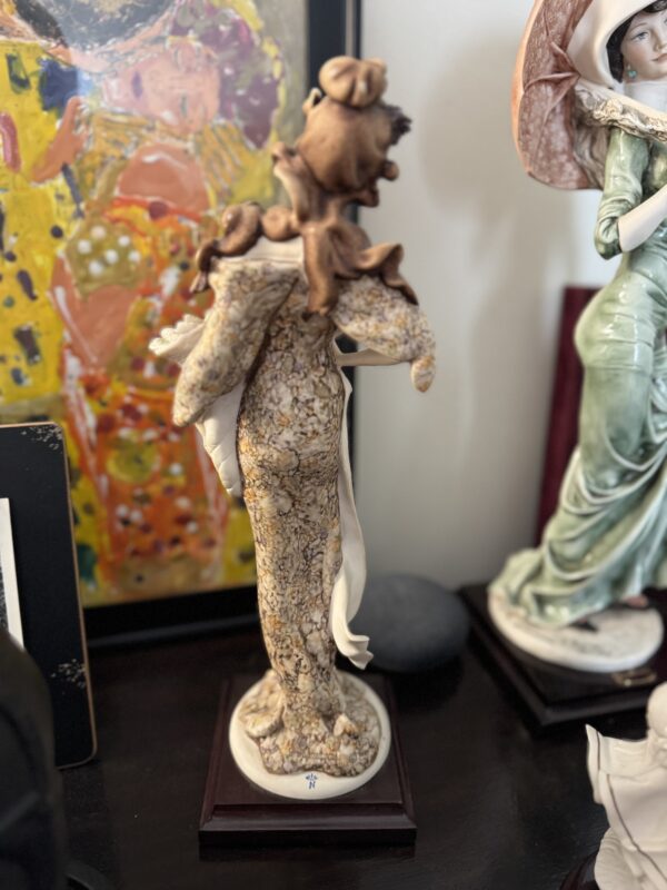 The Armani Lady with Strawberries Figurine depicts a woman in an ornate brown and cream gown, seen from behind with an elaborate updo and white shawl, standing on a wooden base against colorful abstract artwork.