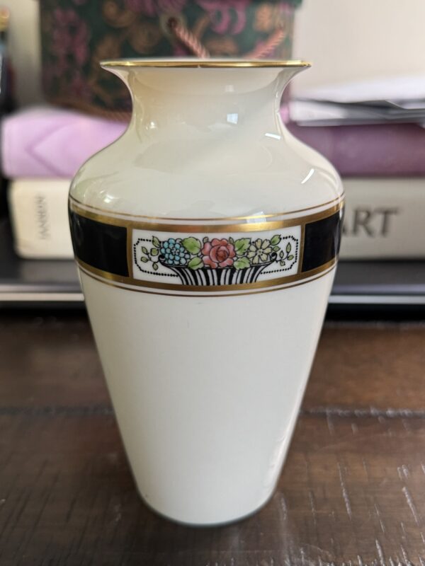 The Lenox Art Deco Vase is a small, white ceramic vase with a narrow neck and flared rim. It features a black band near the top and a colorful rectangular floral basket motif, inspired by classic Lenox style.