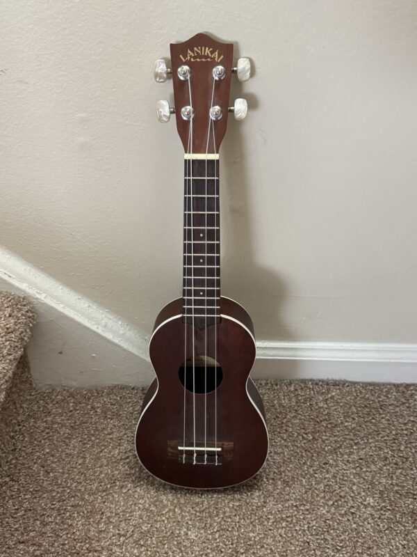 A Lanikai LU 21 ukulele with white tuning pegs leans upright against a beige wall and carpeted stairs, resting on a brown carpet. It has four strings and simple fretboard inlays.