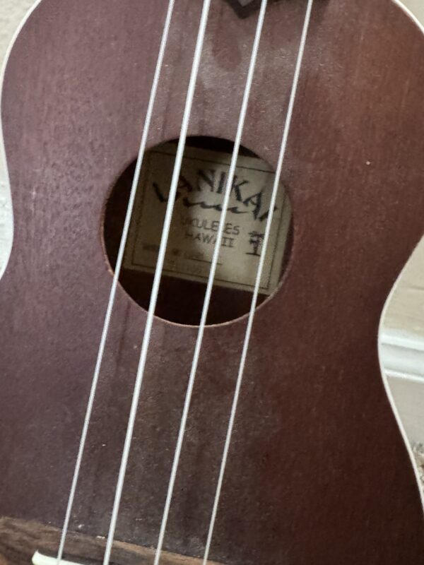 A close-up of the Lanikai LU 21 ukulele highlights its brown body, sound hole, and four white strings. The label inside reads "Lanikai Ukuleles Hawaii." The instrument is set against a light-colored wall.