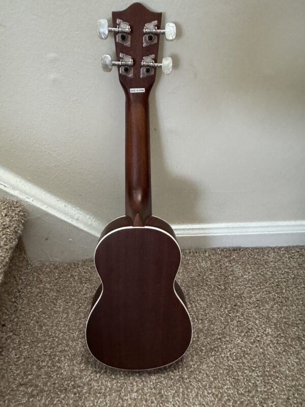 A dark brown Lanikai LU 21 ukulele with a smooth wooden back and white trim stands upright on beige carpet by a light wall and carpeted stairs, showing its tuning pegs and headstock at the top.