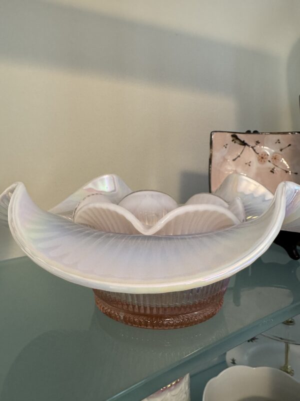 A Fenton Pink Opalescent Bowl with an iridescent ruffled rim sits on a glass shelf. Behind it is a square plate featuring cherry blossoms and several small white dishes from the Fenton Pink Opalescent Bowl Set.