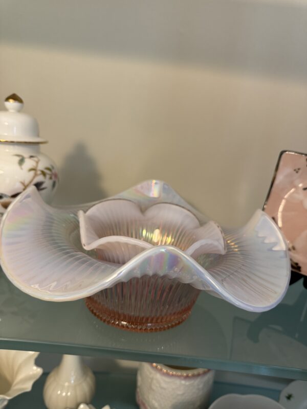 The Fenton Pink Opalescent Bowl Set, with its iridescent sheen, ruffled edges, and ribbed design, is displayed on a glass shelf amid porcelain and glass decor against a neutral background.