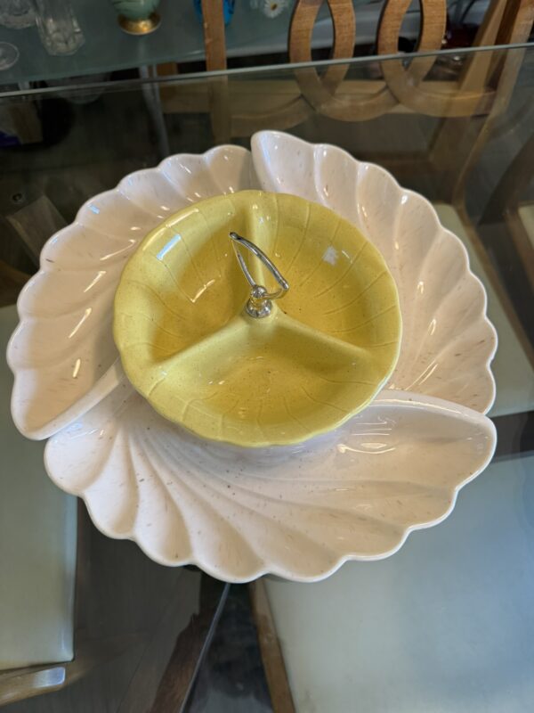 A Lane & Co. MCM Serving Set with three white, leaf-shaped sections sits on a glass table, topped by a yellow, leaf-patterned bowl with a metal handle. Wooden chairs can be seen in the background.