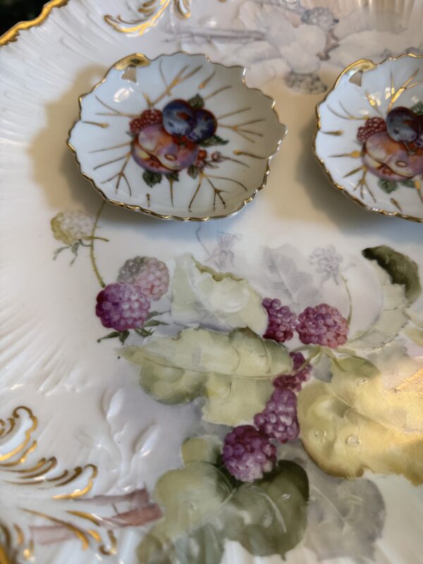 Close-up of two small, scalloped bowls with gold edges and fruit motifs atop a hand-painted M. Redon Plate c. Late 1800s, featuring detailed raspberry and leaf artwork.