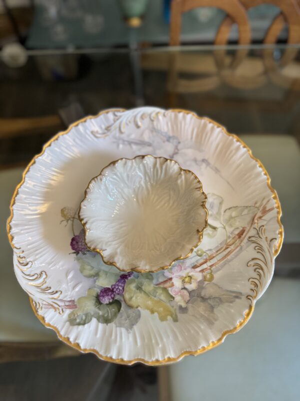 A hand-painted M. Redon plate from the late 1800s, adorned with grapes and florals, is stacked beneath a smaller plain plate with a similar gold-trimmed rim.