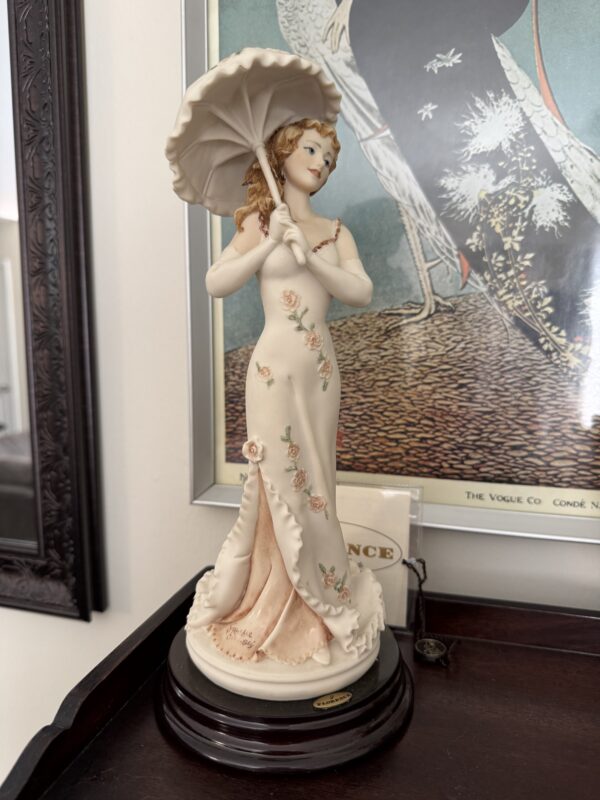The Armani Flower Dress Figurine features a woman in a long floral dress holding a parasol, standing on a dark round base with wavy hair and a gentle smile. Behind her, framed artwork of birds in muted tones decorates the wall.