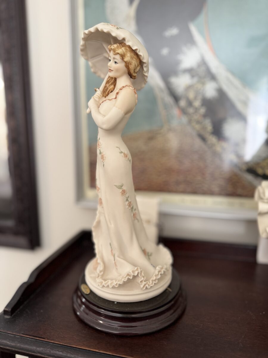 The Armani Flower Dress Figurine depicts a woman in a cream flower dress holding a parasol, with wavy light brown hair, standing on a dark wooden base. Blurred framed artwork and a dark wooden table appear in the background.
