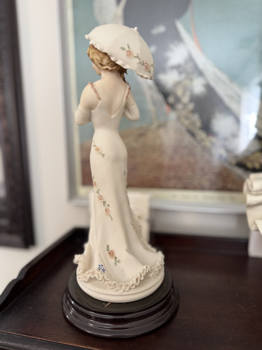 The Armani Flower Dress Figurine features a woman in a cream flower dress and wide-brimmed hat on a dark wooden base, with detailed embroidery and ruffles on her back, set against a softly blurred vintage painting background.
