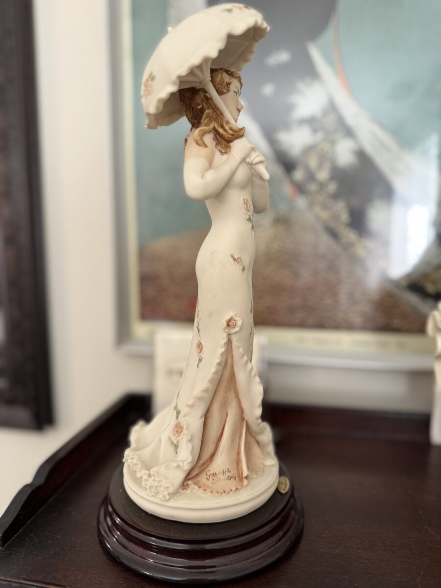 The Armani Flower Dress Figurine depicts a woman in a long floral gown on a dark round base, holding a white parasol. Her brown hair is partly covered by a hat and she looks to the side. A blurred framed artwork appears in the background.
