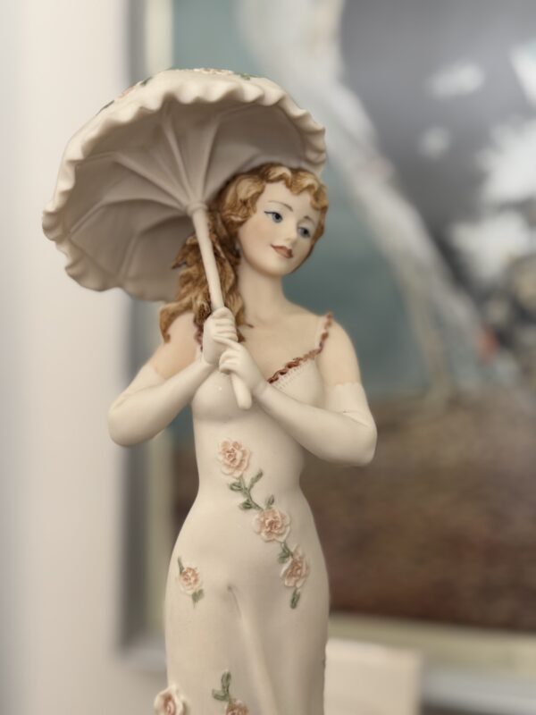 The Armani Flower Dress Figurine features a woman with wavy brown hair holding a white parasol, wearing a sleeveless cream dress with pink floral patterns. A softly blurred background highlights her elegant form.