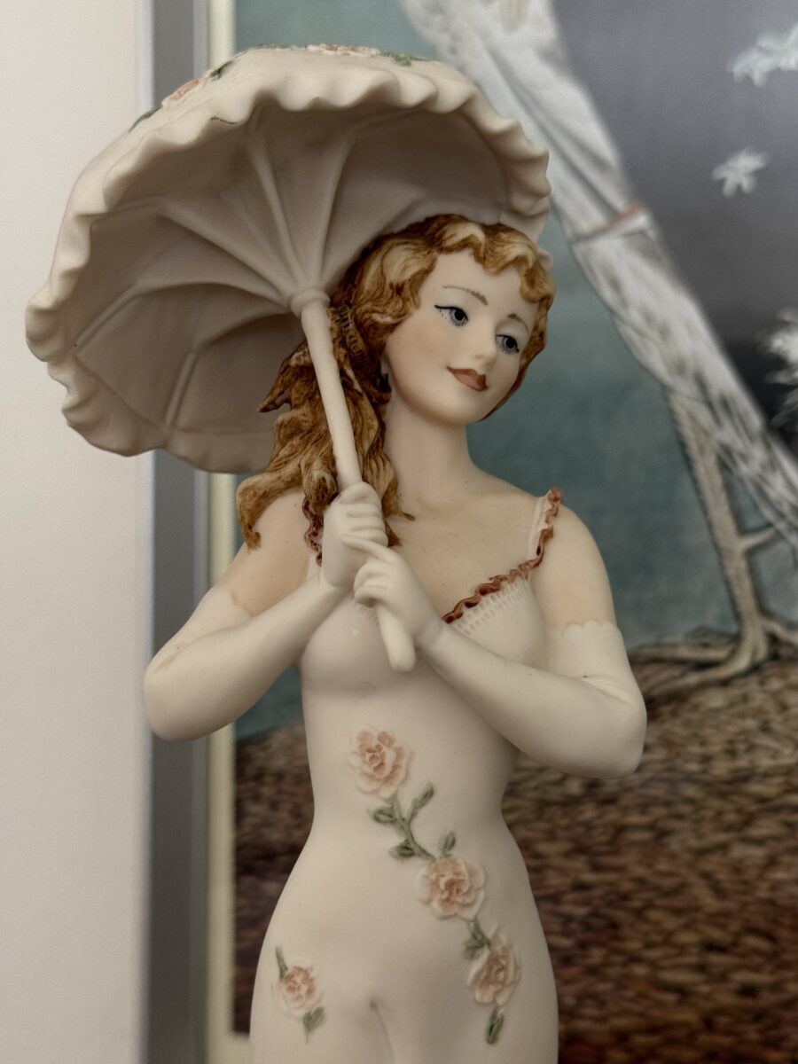 The Armani Flower Dress Figurine depicts a smiling woman with wavy blonde hair, wearing gloves and a sleeveless floral dress adorned with pink roses, holding a large parasol.