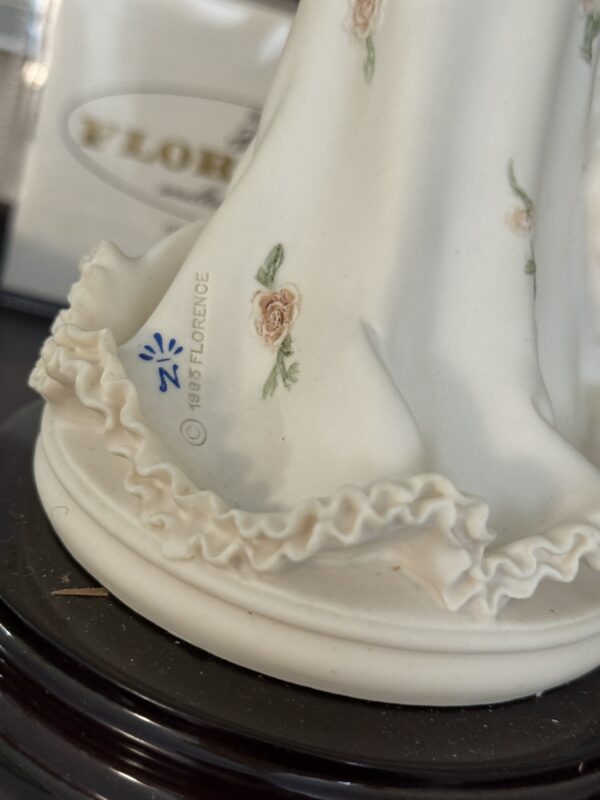 Close-up of the Armani Flower Dress Figurine's ruffled porcelain hem with pastel pink roses. The round white base features the 1985 FLORENCE marking and blue logo. Background is softly blurred.