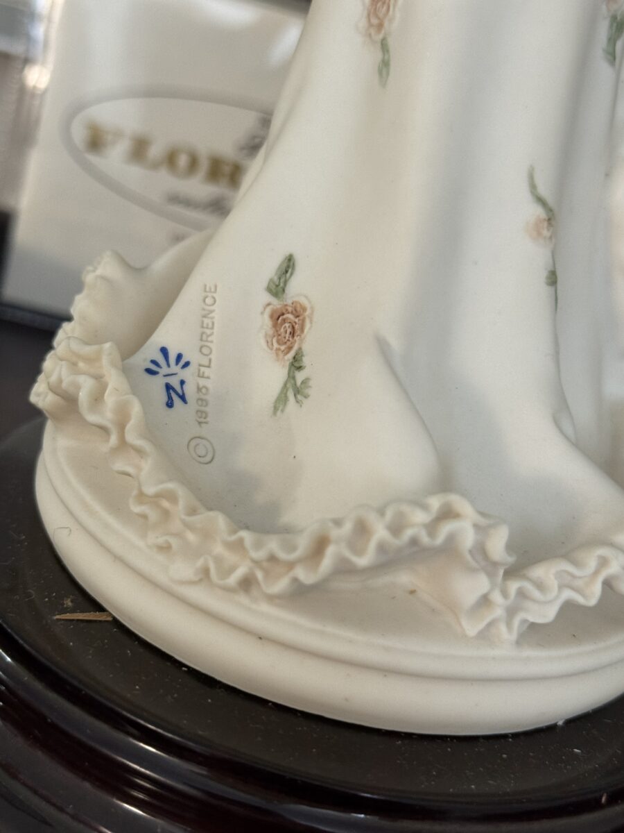 Close-up of the Armani Flower Dress Figurine's ruffled porcelain hem with pastel pink roses. The round white base features the 1985 FLORENCE marking and blue logo. Background is softly blurred.
