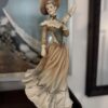 The Armani Vanessa Figurine showcases a woman in a vintage beige dress with green accents and a wide-brimmed hat, holding a fan. She stands on a dark base labeled "FLORENCE," with a framed painting seen in the background.