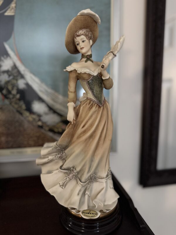 The Armani Vanessa Figurine showcases a woman in a vintage beige dress with green accents and a wide-brimmed hat, holding a fan. She stands on a dark base labeled "FLORENCE," with a framed painting seen in the background.