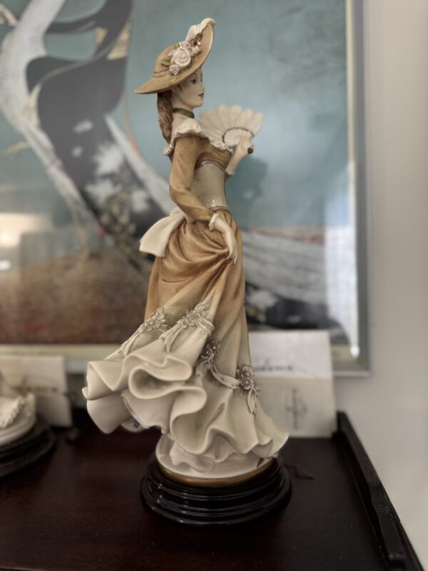 The Armani Vanessa Figurine, made of porcelain, features a Victorian-era woman in a flowing tan and cream dress with an Armani-style floral hat, holding a fan on a black base. A painting and white envelope are softly blurred in the background.