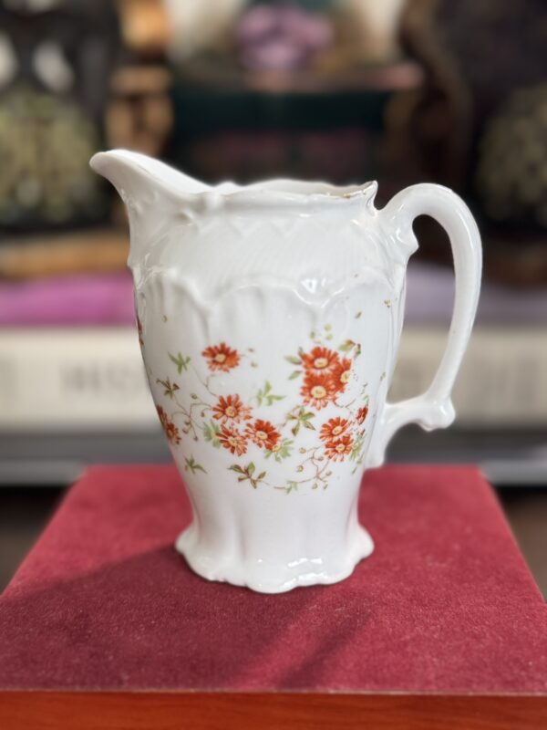 The Floral Creamer is a small, ornate white porcelain piece with a curved handle and spout, adorned with orange-red flowers and green leaves. It sits on red velvet, its intricate details highlighted by a softly blurred background.