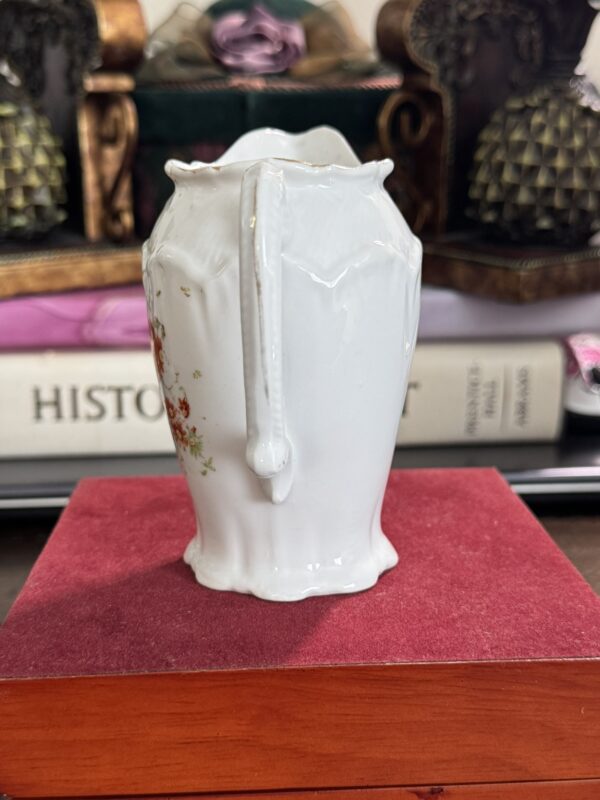 The Floral Creamer, made of white porcelain with a curved handle, floral side design, and scalloped edges, rests on a red velvet box. Behind it are books—including one titled HISTORY—and two lattice-patterned decor items.