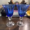 Two Vintage Blue Glass Wine Glasses with clear stems rest on a dark wooden table. The cobalt glass has tiny bubbles, while the blurred background shows decorative objects, including figurines and a stacked book.