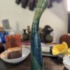 The Vintage Ceramic Art Studio Vase, tall and narrow with a glossy green-blue ombre finish, curved tilted neck, and small black feet, stands on a wooden table against a backdrop of blurred colorful decor.