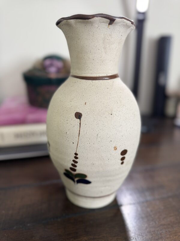 A cream-colored Tonala Pottery Vase from Mexico features brown and green abstract leaf and dot patterns, a rounded body, and a flared uneven rim. It is displayed on a wooden surface with blurred background objects like a book and container.