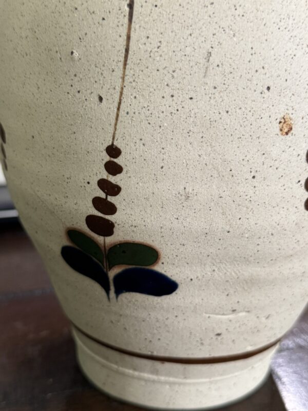 A close-up of the Tonala Pottery Vase Mexico, beige and speckled with a painted brown vertical line, clustered ovals, and stylized green and dark blue leaves, sits on a dark wooden surface. Inspired by traditional Mexican design.