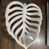 A cream-colored ceramic tray shaped like a monstera leaf with large cutouts mimicking the leaf’s natural holes sits on a dark wooden surface. The trays glossy finish reflects light, emphasizing its smooth texture and organic form.