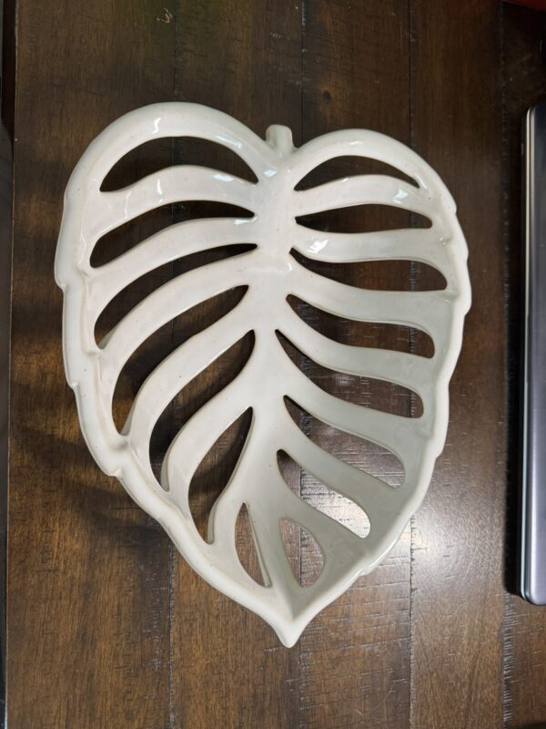 A cream-colored ceramic tray shaped like a monstera leaf with large cutouts mimicking the leaf’s natural holes sits on a dark wooden surface. The trays glossy finish reflects light, emphasizing its smooth texture and organic form.